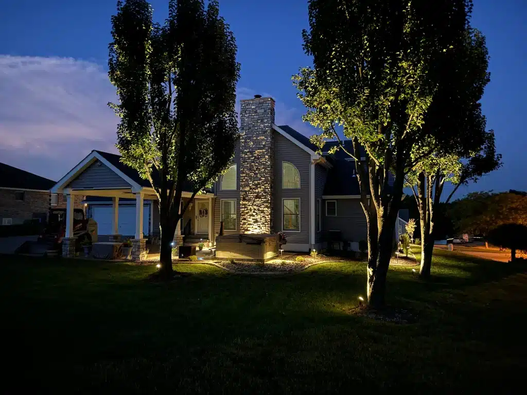 Professional LED landscape lighting installation by Action Landscaping in St. Louis, featuring illuminated trees and a stone chimney at twilight.